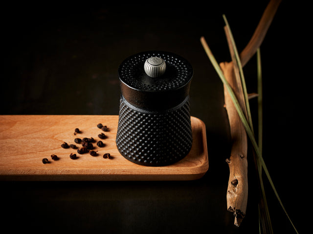 Pfeffermühle BALI aus Gusseisen in schwarz von Peugeot auf einem Holzbrett mit Pfefferkörnern